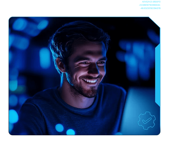 Homem branco de cabelo e barba castanhos, usa camiseta escura, em ambiente escuro olhando para uma tela e sorrindo